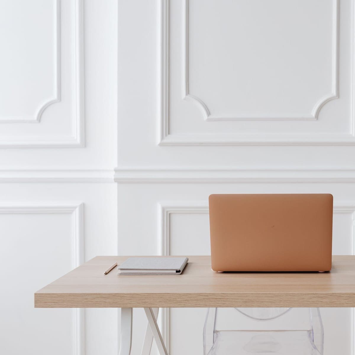 A laptop and notebook on a light wood desk beside a small vase of flowers.
