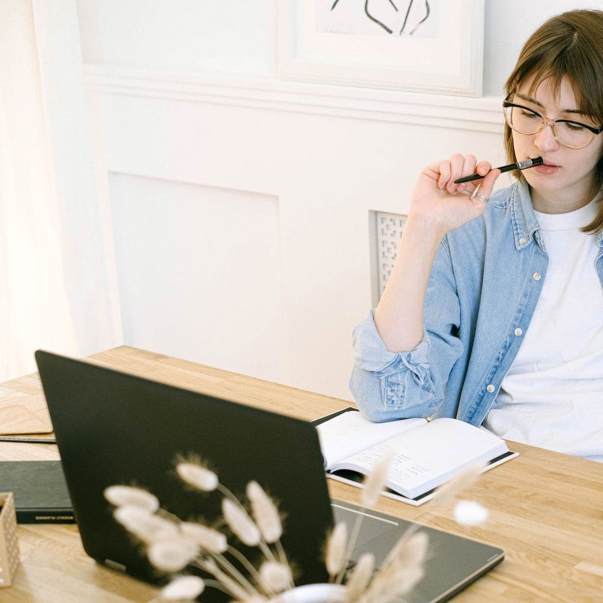 A woman at a bright desk, pen to her chin, thinking over an open laptop.